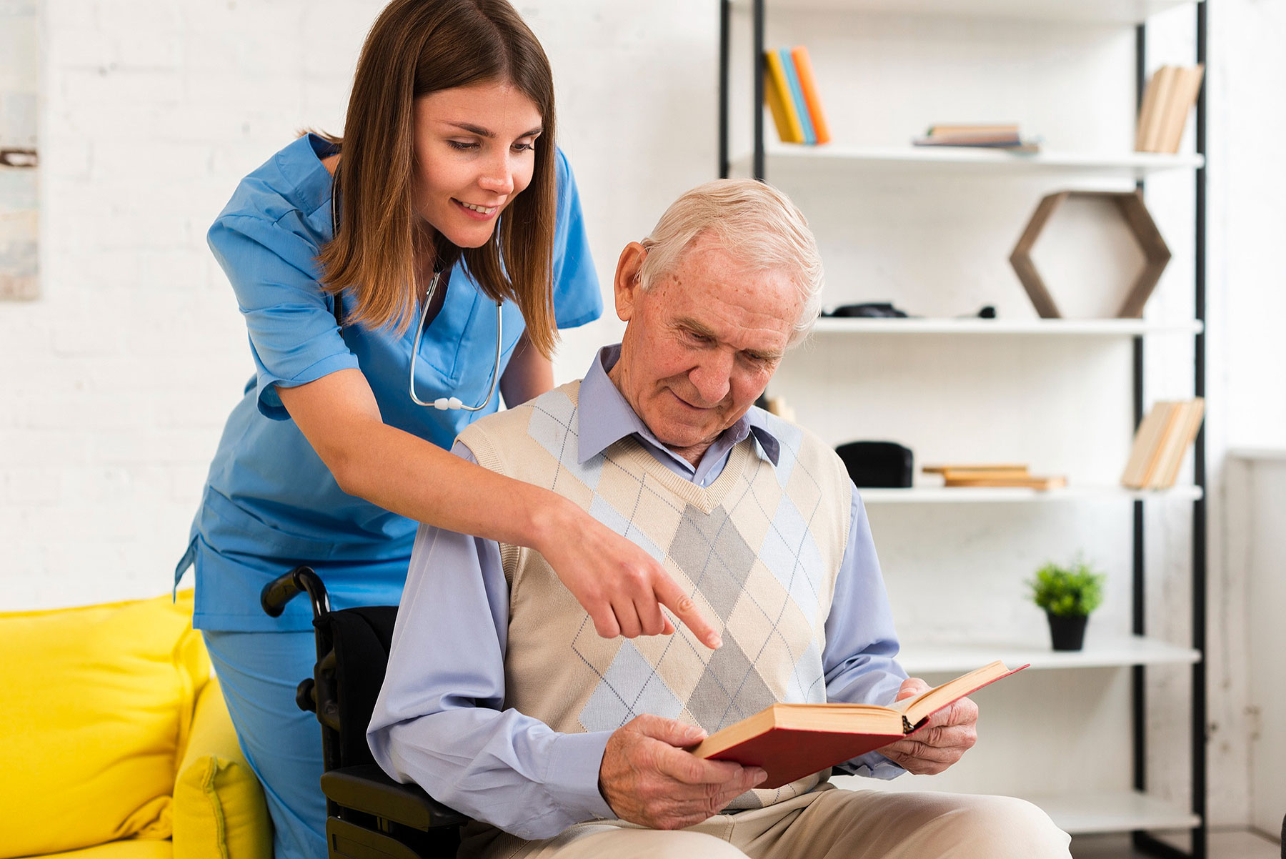 A compassionate care caregiver wearing a blue uniform pointing at a book. An elderly man reading with a smile