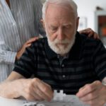 A gentleman dealing with dementia working on a puzzle, with the hands of his wife on his back supporting him
