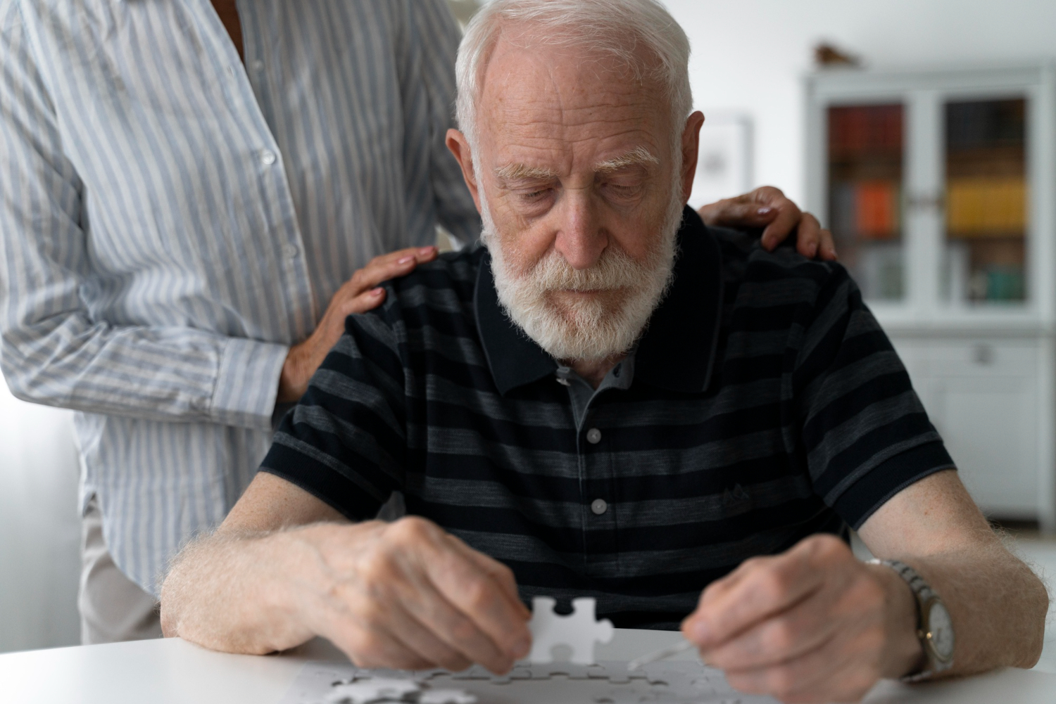 A gentleman dealing with dementia working on a puzzle, with the hands of his wife on his back supporting him